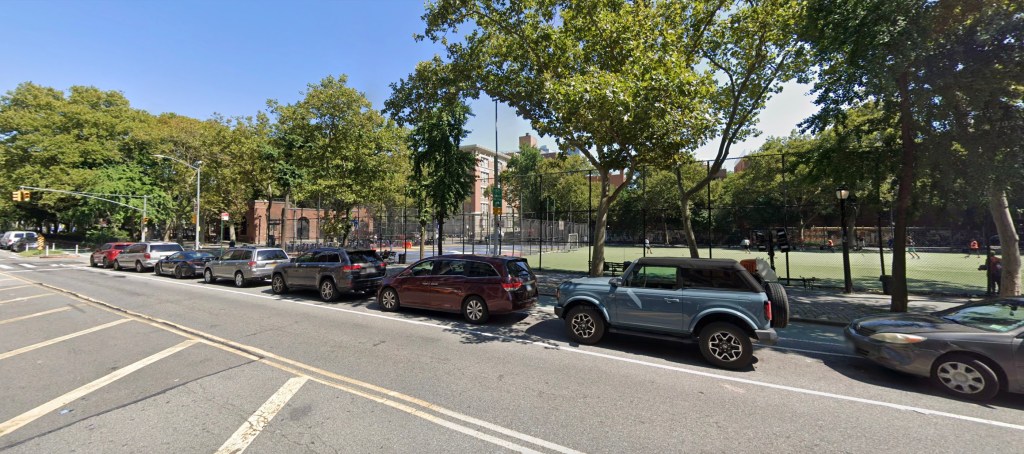 Street scene with parked cars lining a road next to a park with a playing field.